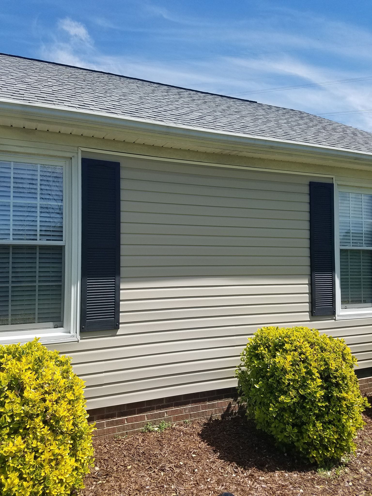 Tan house with white-framed windows, black shutters, and small yellow-green bushes in front.