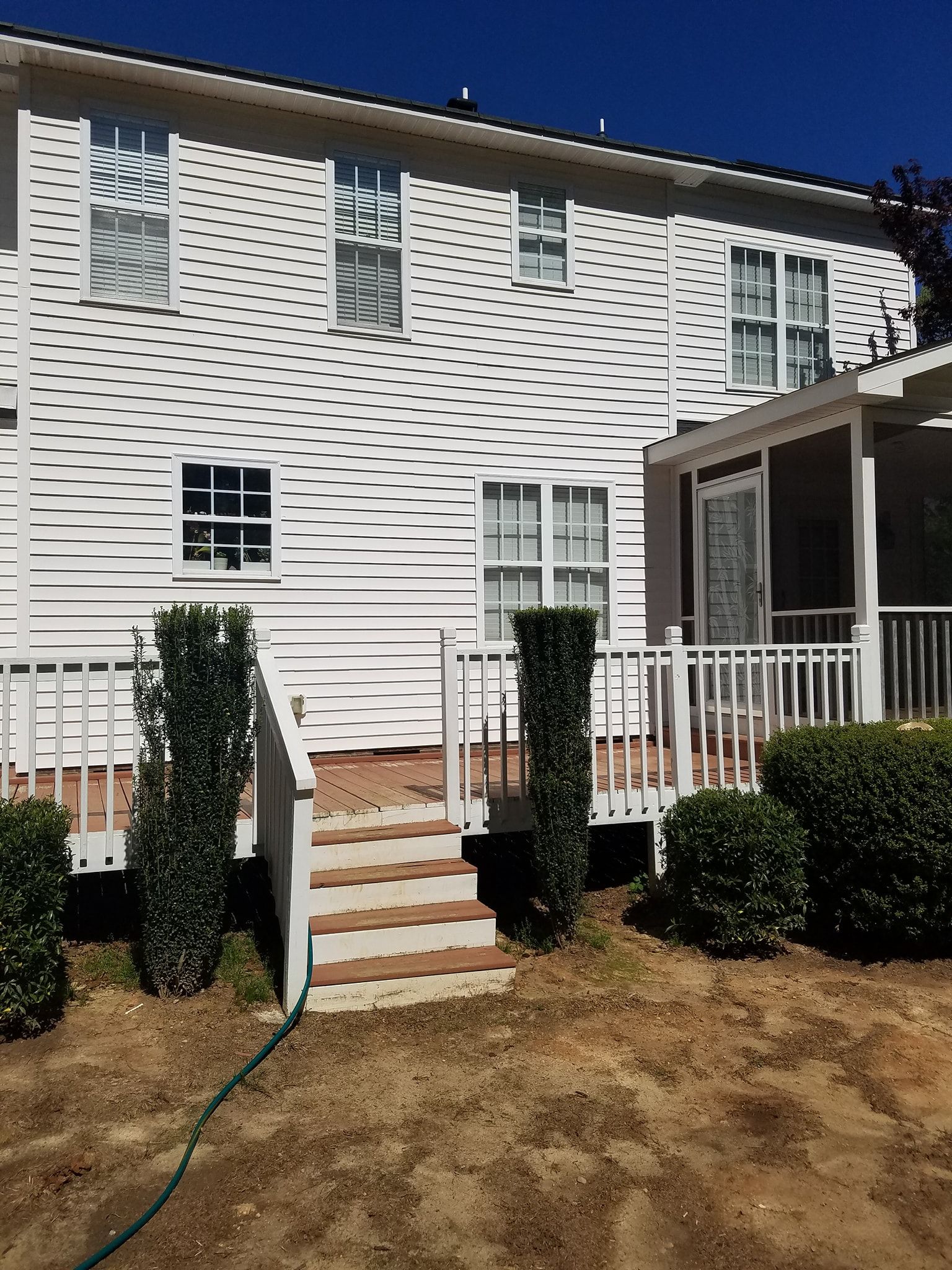 Back of a white house with a deck and steps, surrounded by landscaping.