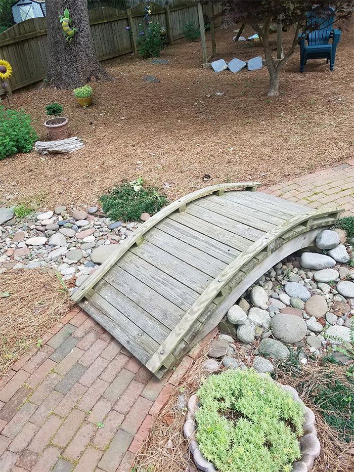Wooden arched bridge over a creek bed in a backyard.
