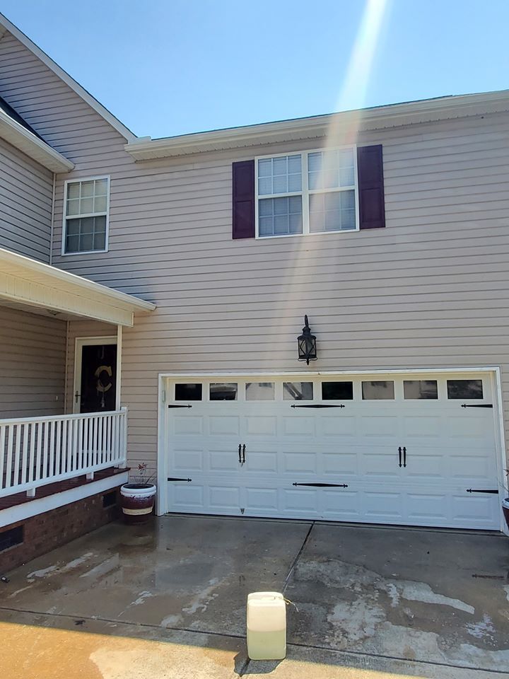 Beige house with white garage door, dark shutters, and porch. Sunlight overhead.