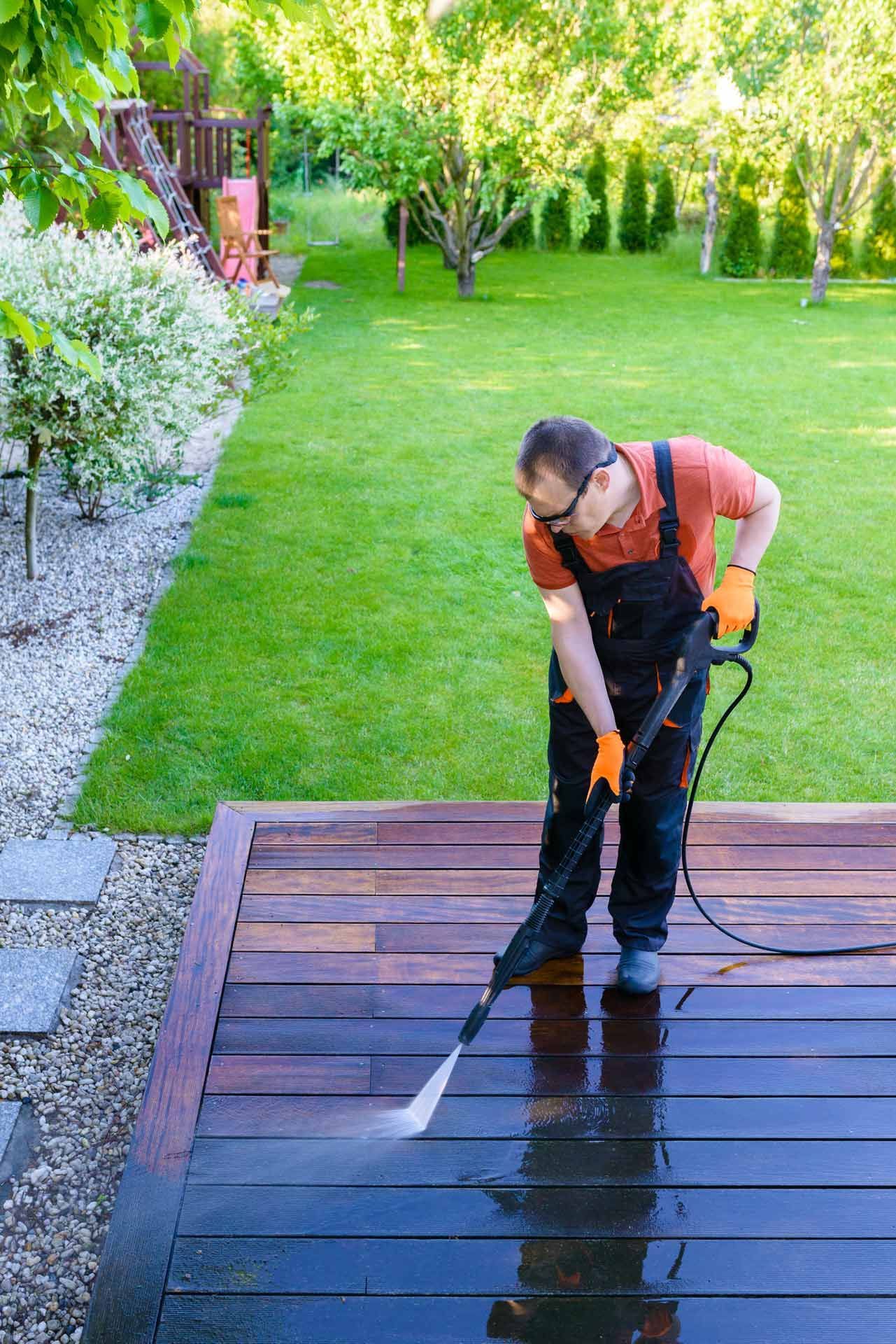 Man in orange shirt and overalls power washing a wooden deck in a backyard