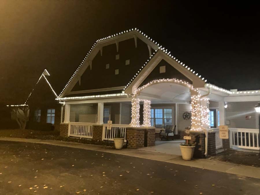 A building at night with Christmas lights on the roof and columns; a porch is visible.