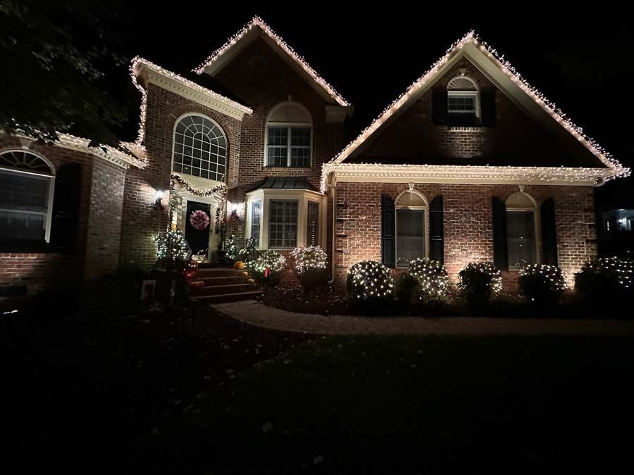 Brick house at night with Christmas lights outlining the roof and bushes, a walkway leads to the front door.