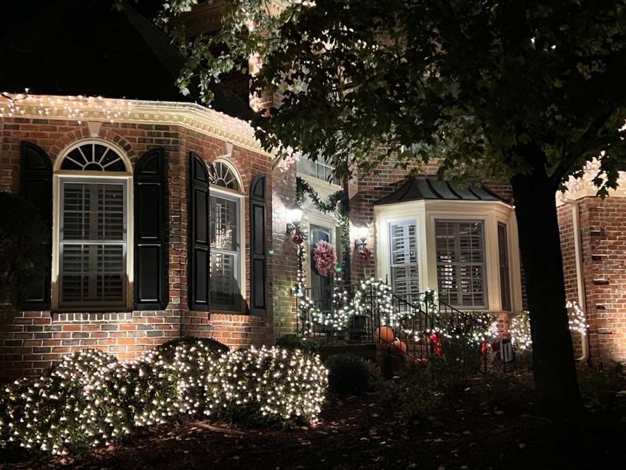 A brick house with Christmas lights and decorations at night; bushes are lit up.