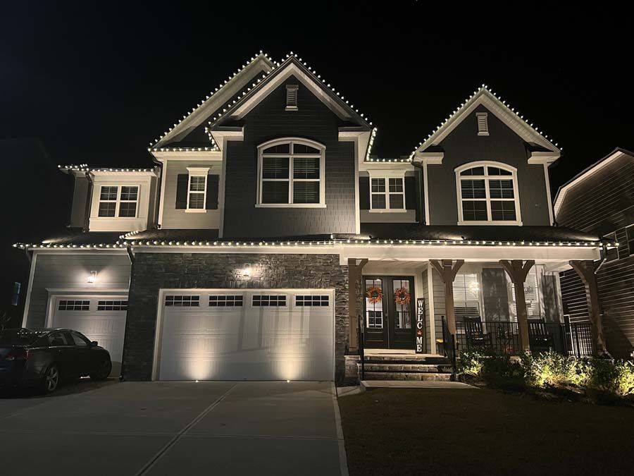 Two-story house at night, lit with white Christmas lights along rooflines and landscaping.