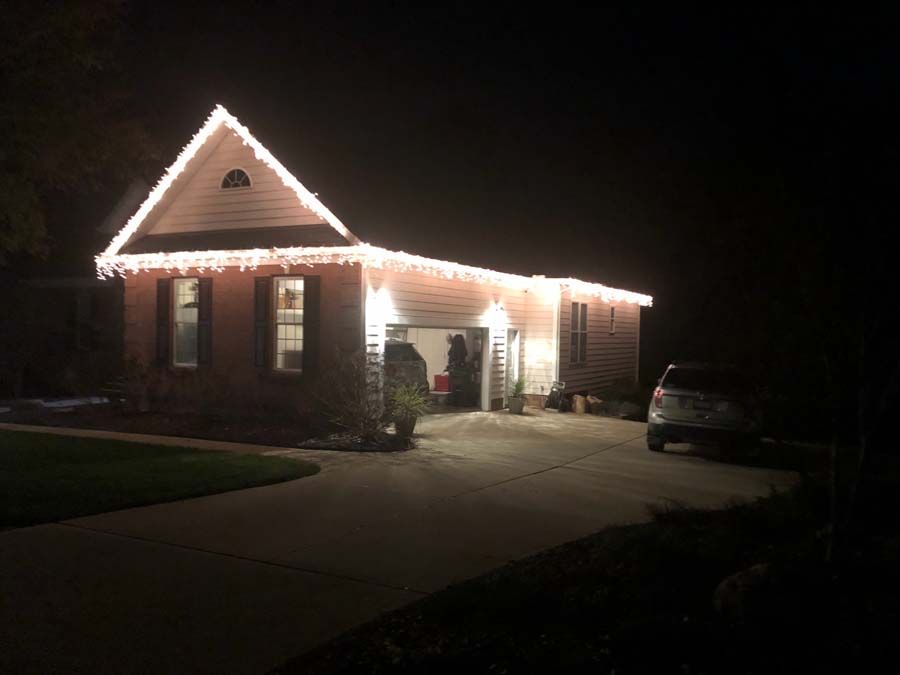 House at night, lit with white Christmas lights along the roofline and above the garage; driveway with car.