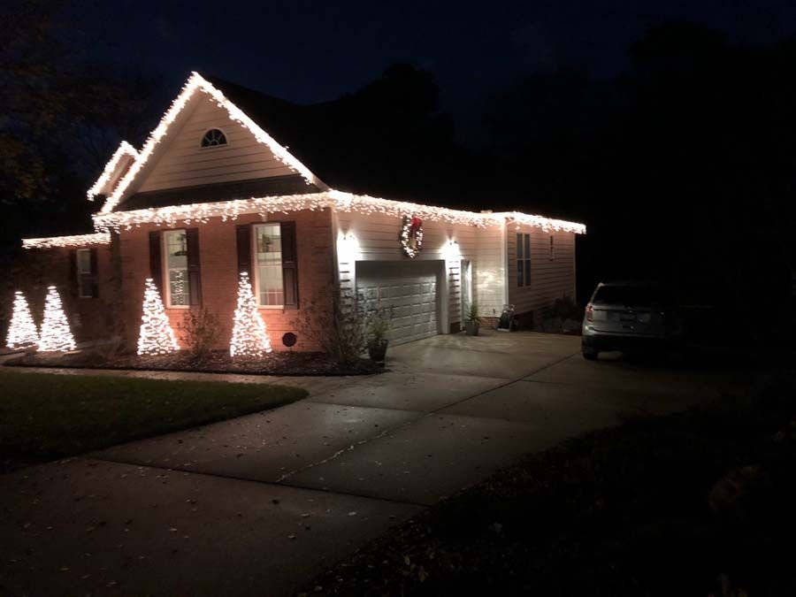 House decorated with white Christmas lights; a driveway, car, and lit decorative trees.