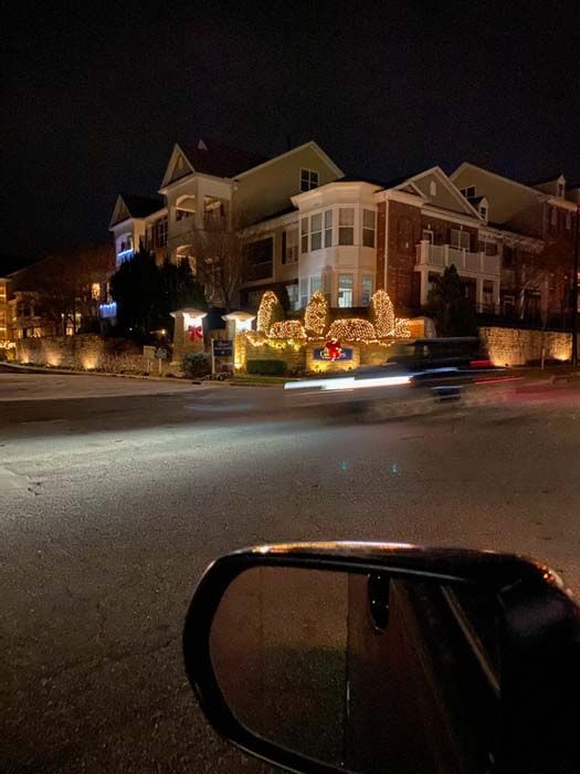 Night scene: buildings with holiday lights, seen from a car. Blurry car headlights illuminate the road and side mirror.