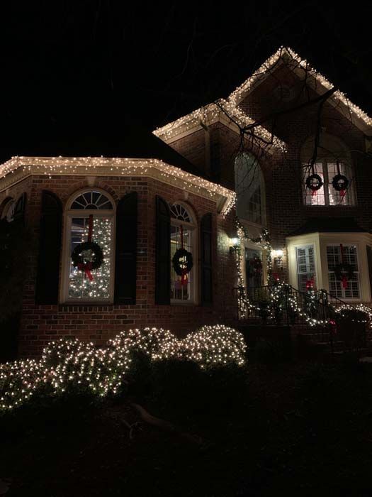 A brick house illuminated with Christmas lights. Wreaths adorn windows, bushes sparkle below.