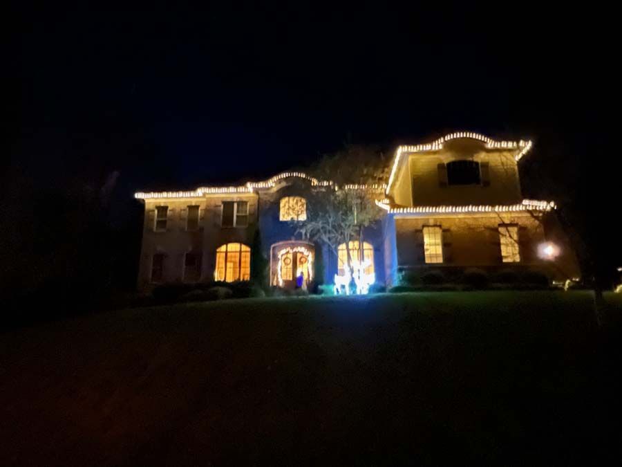 Large house at night, lit with white Christmas lights along the roofline and around the windows; dark sky.