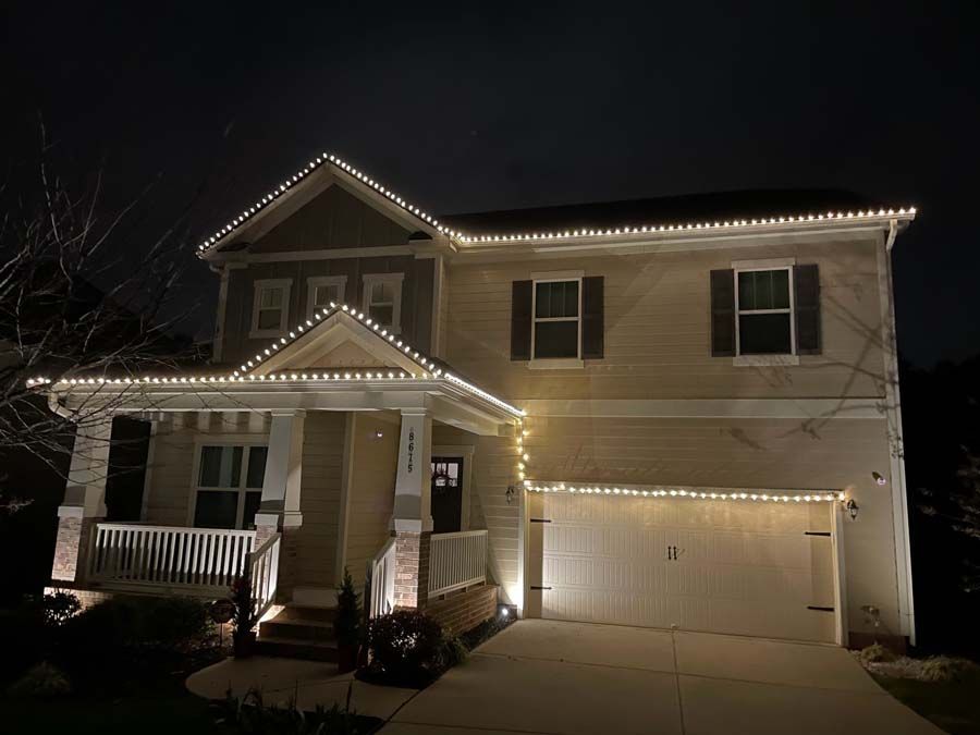 Two-story house at night illuminated by white string lights along the roof and garage, with spotlights.