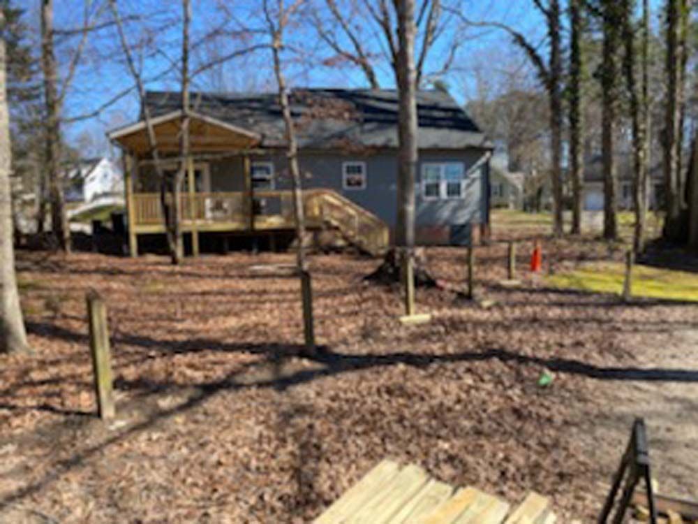 A small gray house with a wooden deck and stairs, surrounded by trees and fallen leaves.
