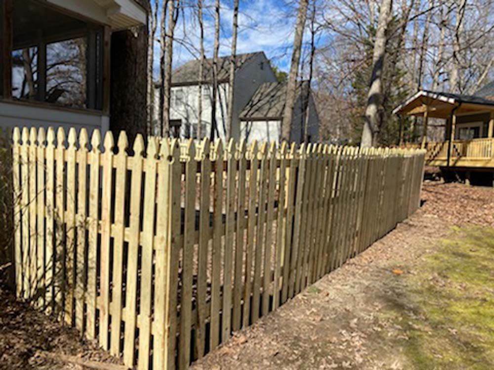 Wooden picket fence in a yard with a house and trees in the background.