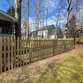 A wooden picket fence surrounds a yard with bare trees, a house, and a sunny sky.
