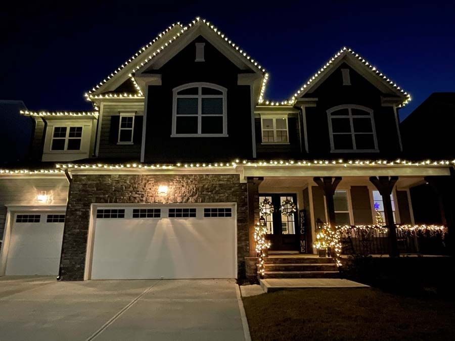 Two-story house with white Christmas lights outlining the roof, windows, and porch at night.