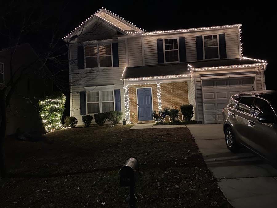 Two-story house at night decorated with white Christmas lights on the roof and bushes.