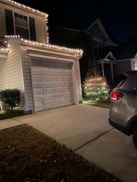 A house decorated with white Christmas lights, with a silver SUV parked in the driveway at night.