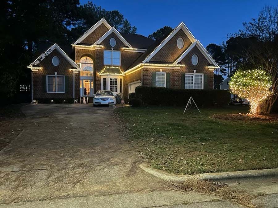 House decorated with Christmas lights at dusk; car in the driveway, tree also lit.