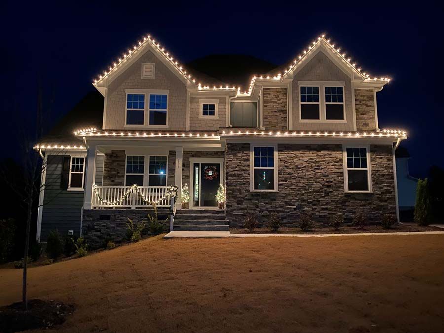 House with white Christmas lights along the roofline at night; stone and siding facade.