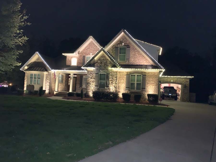 Nighttime view of a brick house illuminated with white lights along the roof and warm floodlights.