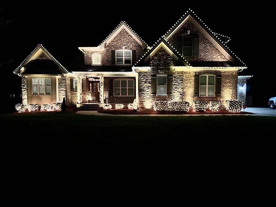 Lit up brick house at night, decorated with white lights around the roof and bushes.