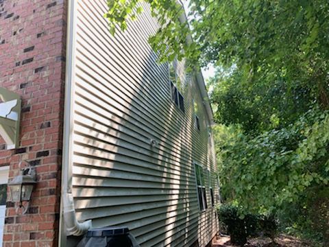 Side of a two-story beige house with siding, red brick, and a rain barrel next to greenery.