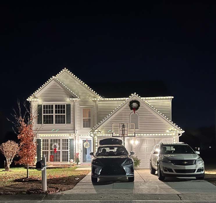 White house at night with Christmas lights, a car in the driveway and another car on the right.