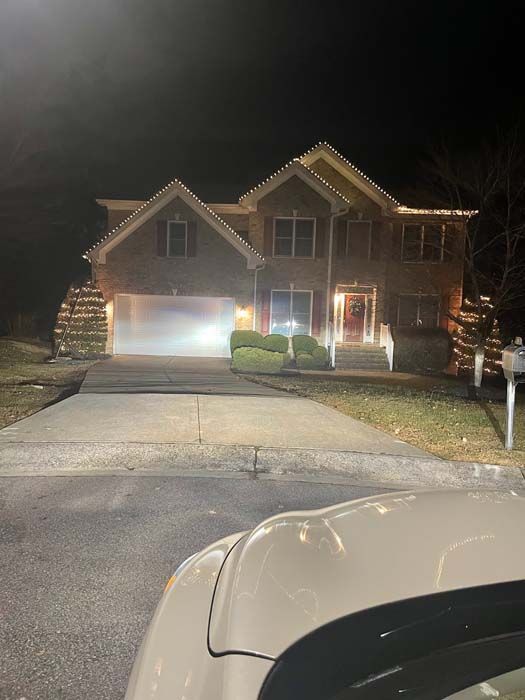 Night view of a house decorated with lights; a car's hood in the foreground, driveway leads to the house.
