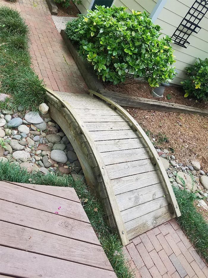 Wooden arched bridge over a stone and brick path, surrounded by greenery.