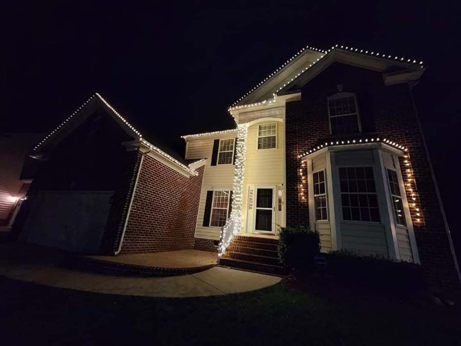 A two-story house at night, decorated with white Christmas lights along roof edges and entryway.