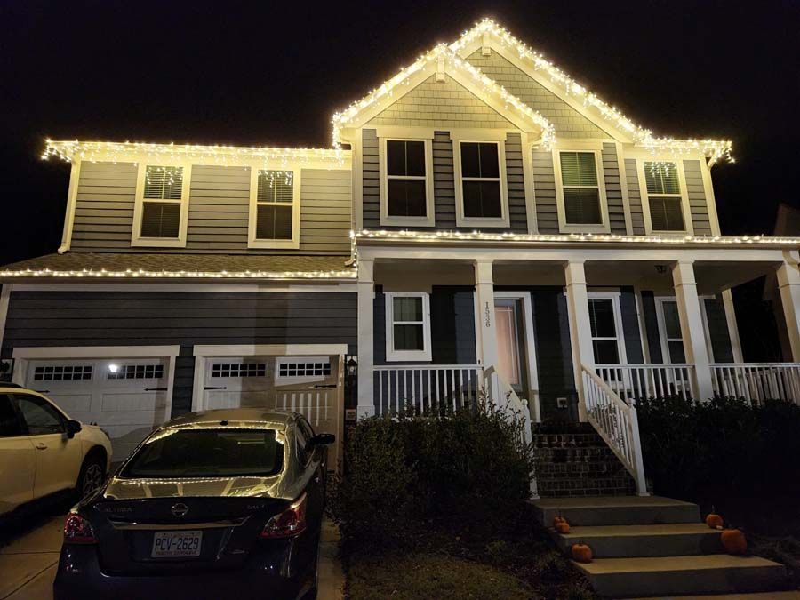 A house at night with yellow holiday lights, a car in the driveway, and pumpkins on the steps.