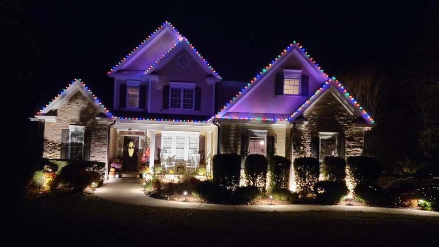 House at night, decorated with multi-colored Christmas lights. Uplighting on shrubs.