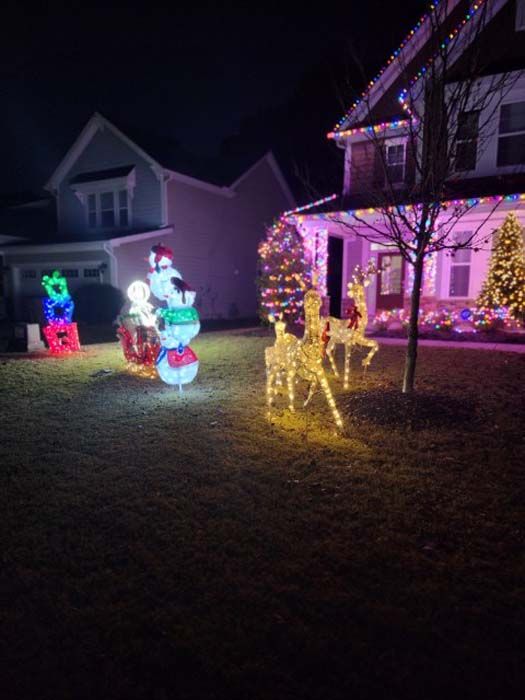 Christmas light display with illuminated reindeer, snowman, and gifts on a lawn. A house with lights is in the background.