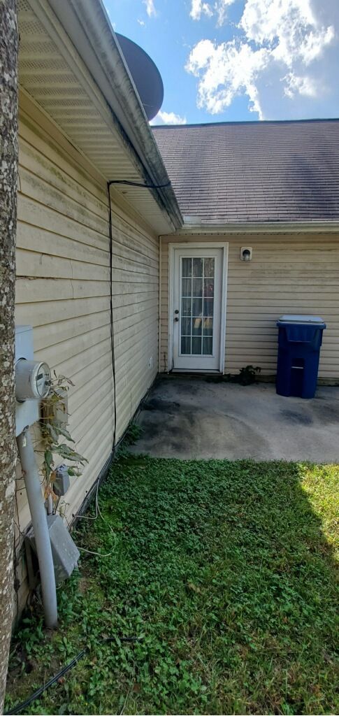 Side of a house with a door, trash can, and overgrown grass along a walkway.