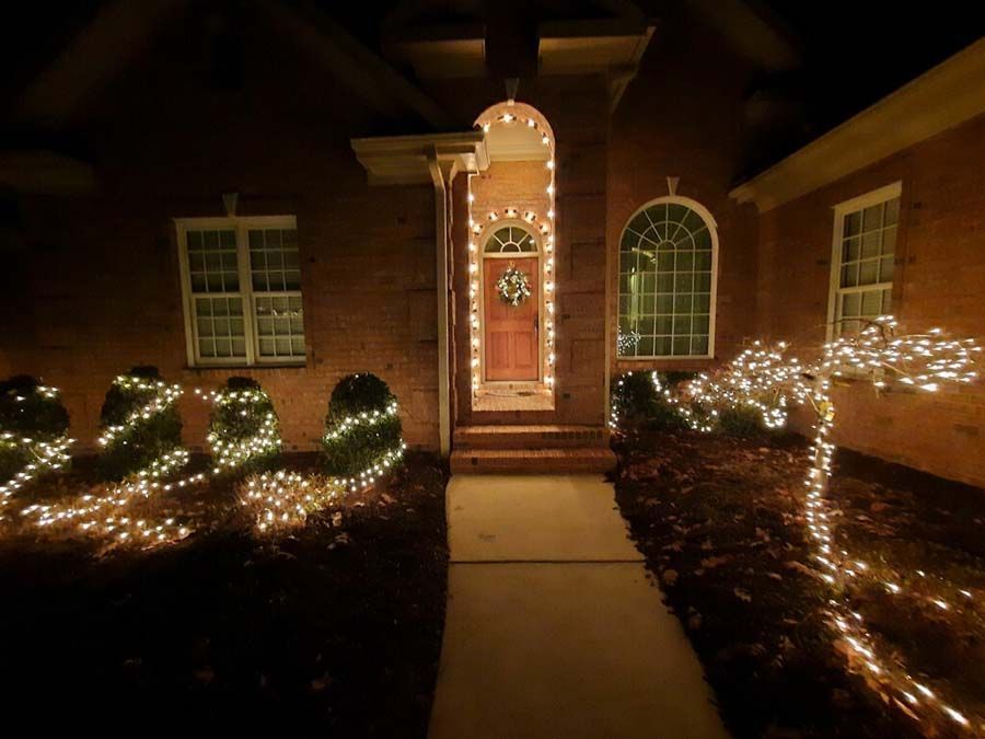 House exterior at night, decorated with white Christmas lights. Brown door, brick facade, bushes outlined with lights.