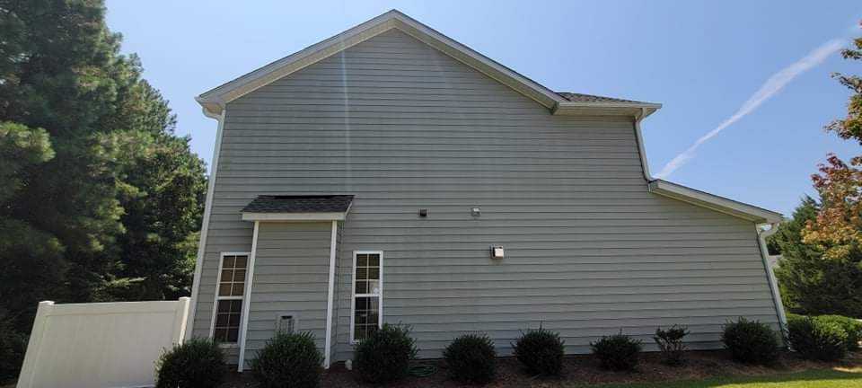 Side view of a two-story gray house with white trim. Lush green trees and bushes surround the home. Bright blue sky.