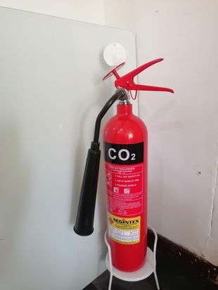 Extintor de CO2 rojo montado en una pared blanca junto a la manija de un tubo vertical negro.