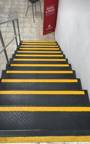 Escalera con peldaños negros y franjas amarillas de seguridad, que desciende entre muros de piedra y una barandilla metálica.