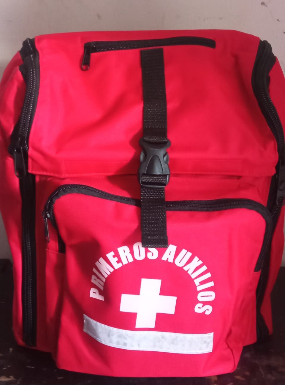 Mochila roja de primeros auxilios con una cruz blanca y el texto “PRIMEROS AUXILIOS”.