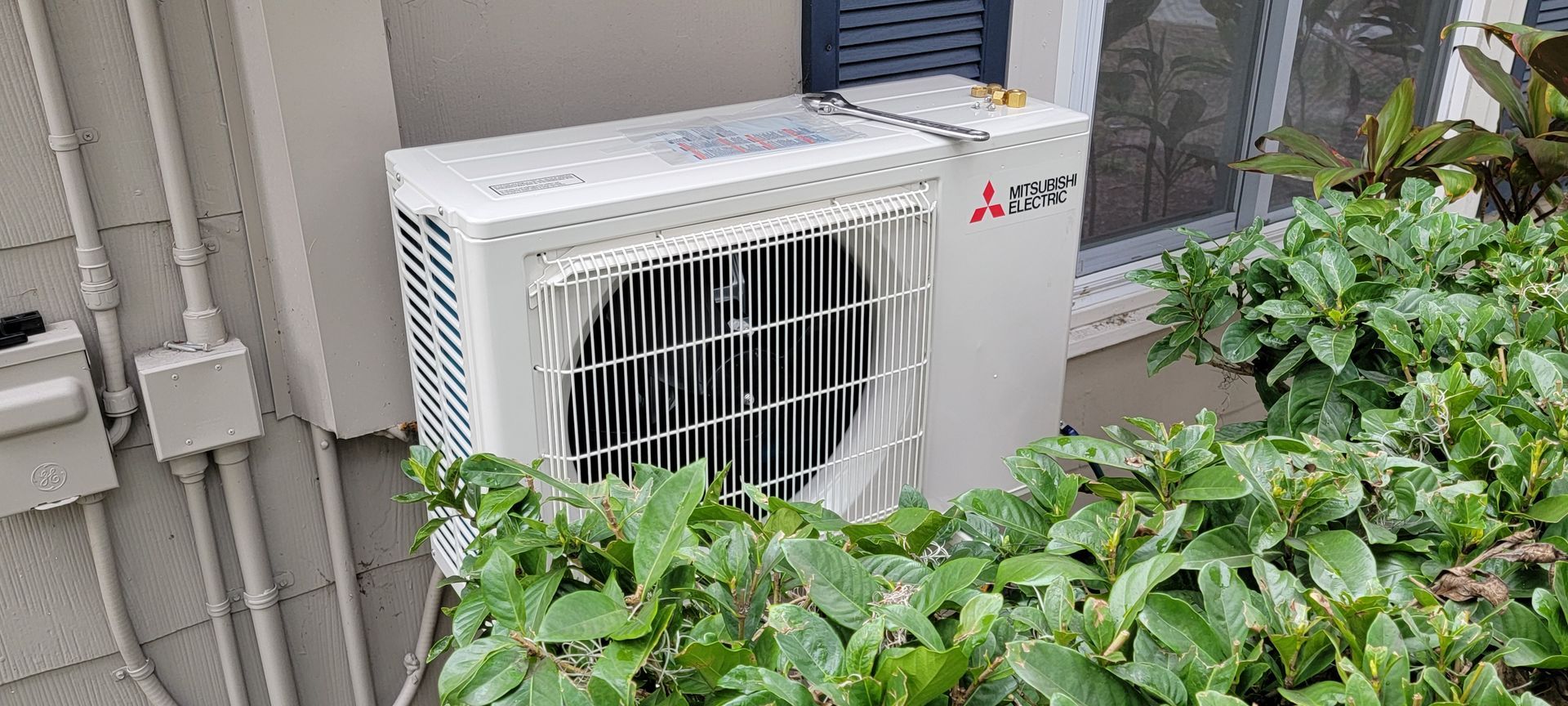 A white air conditioner is sitting on the side of a building next to a bush.