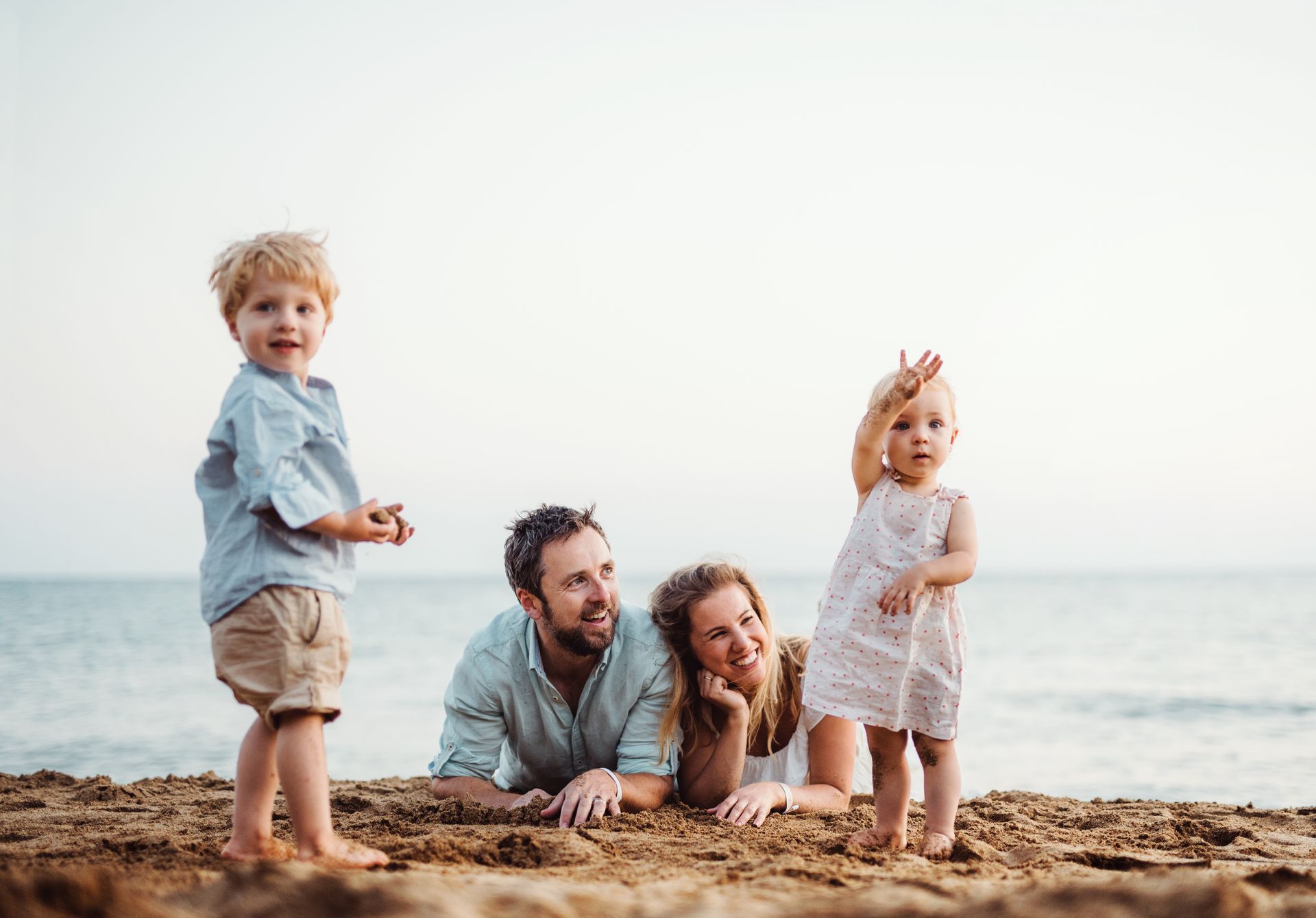 Family of four at the beach; parents lying down, two young children standing, one pointing at the sea.