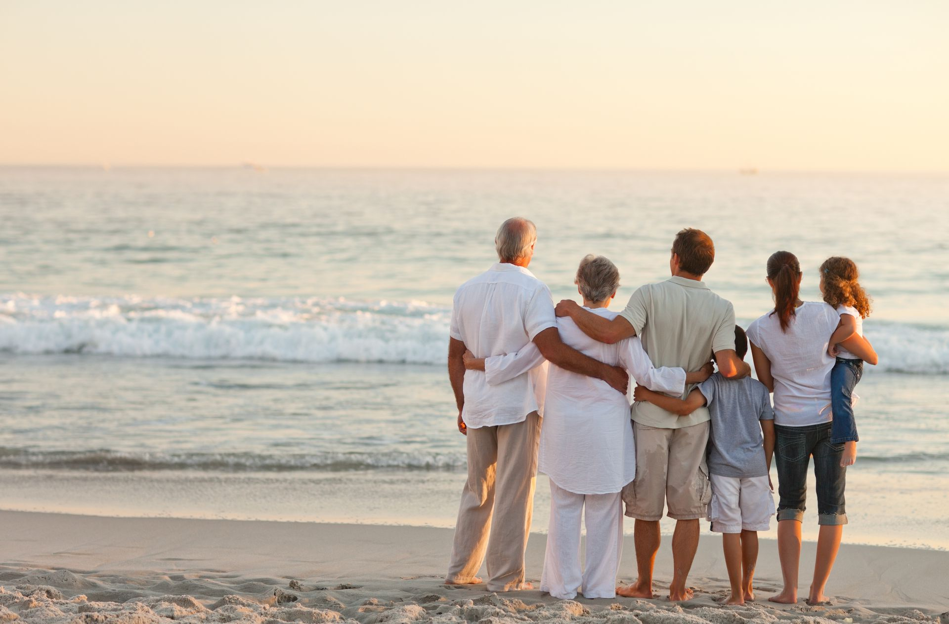 Family embraces on beach, watching ocean at sunset.