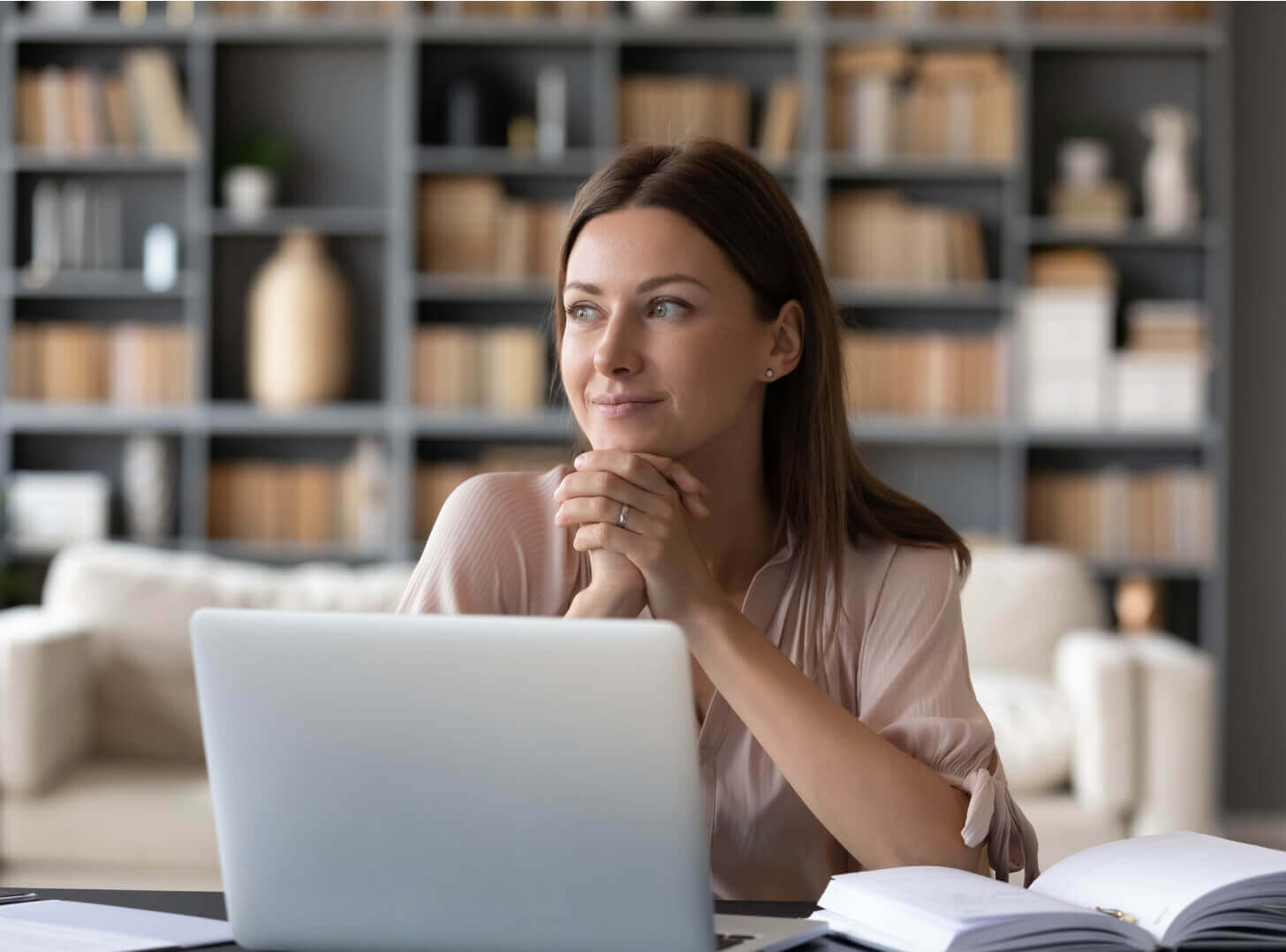 Woman at laptop, resting chin on hands, looking thoughtfully toward the distance. Bookshelves in background.