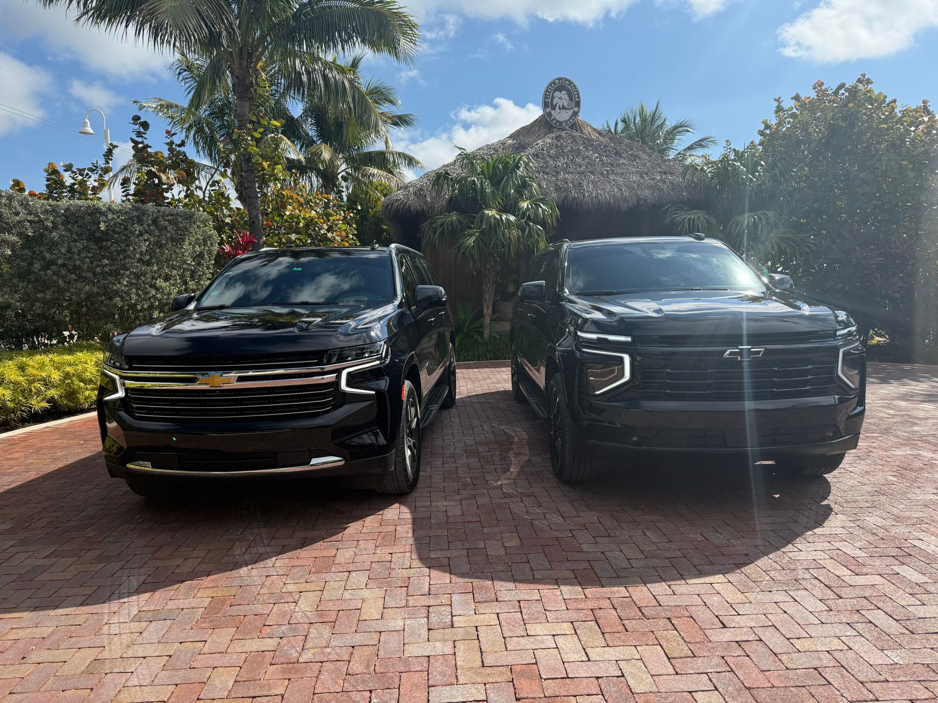Two black Chevrolet trucks parked on a brick driveway; palm trees and a tiki hut in the background.