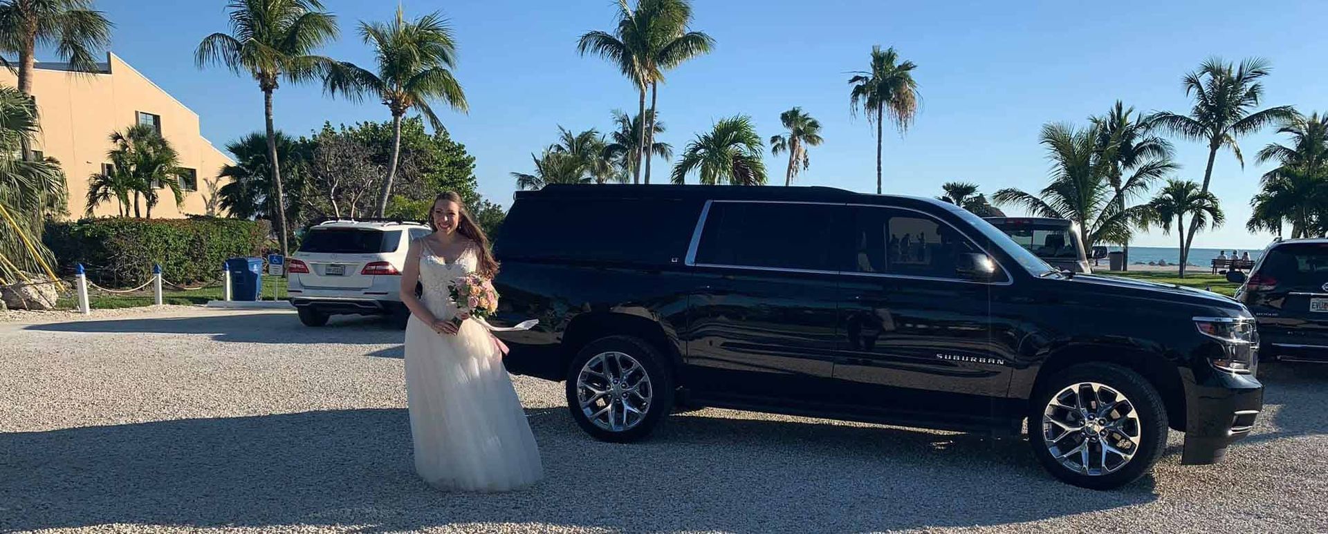 Bride in white dress with bouquet next to black SUV; beach setting.