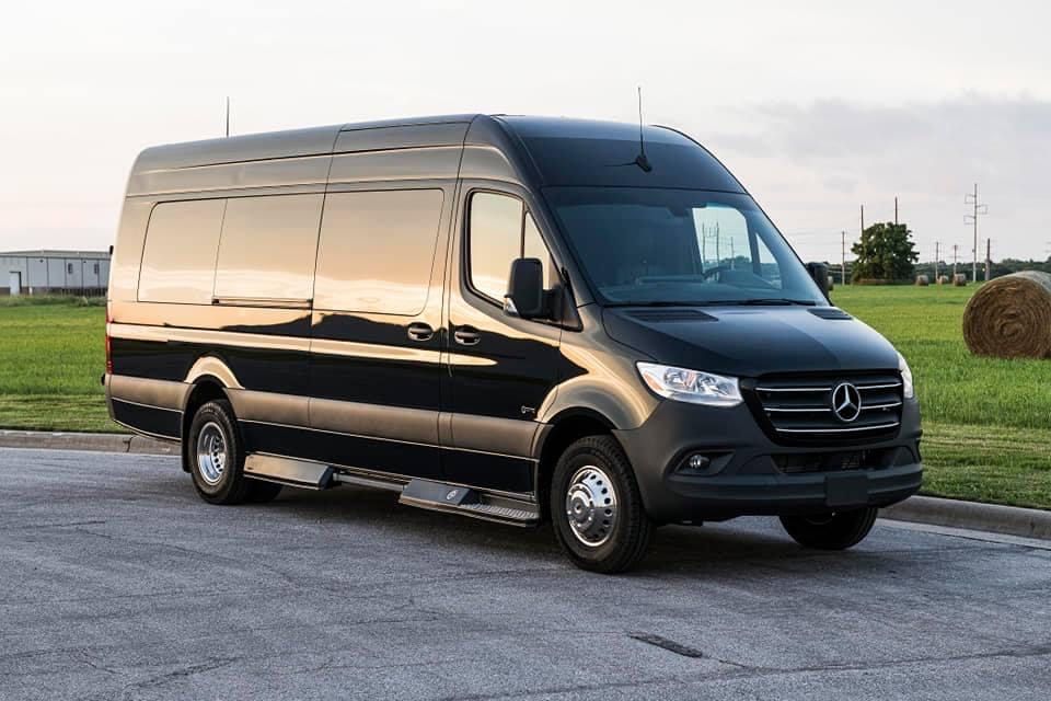 Black Mercedes-Benz Sprinter van parked on asphalt, field with hay bale in the background.