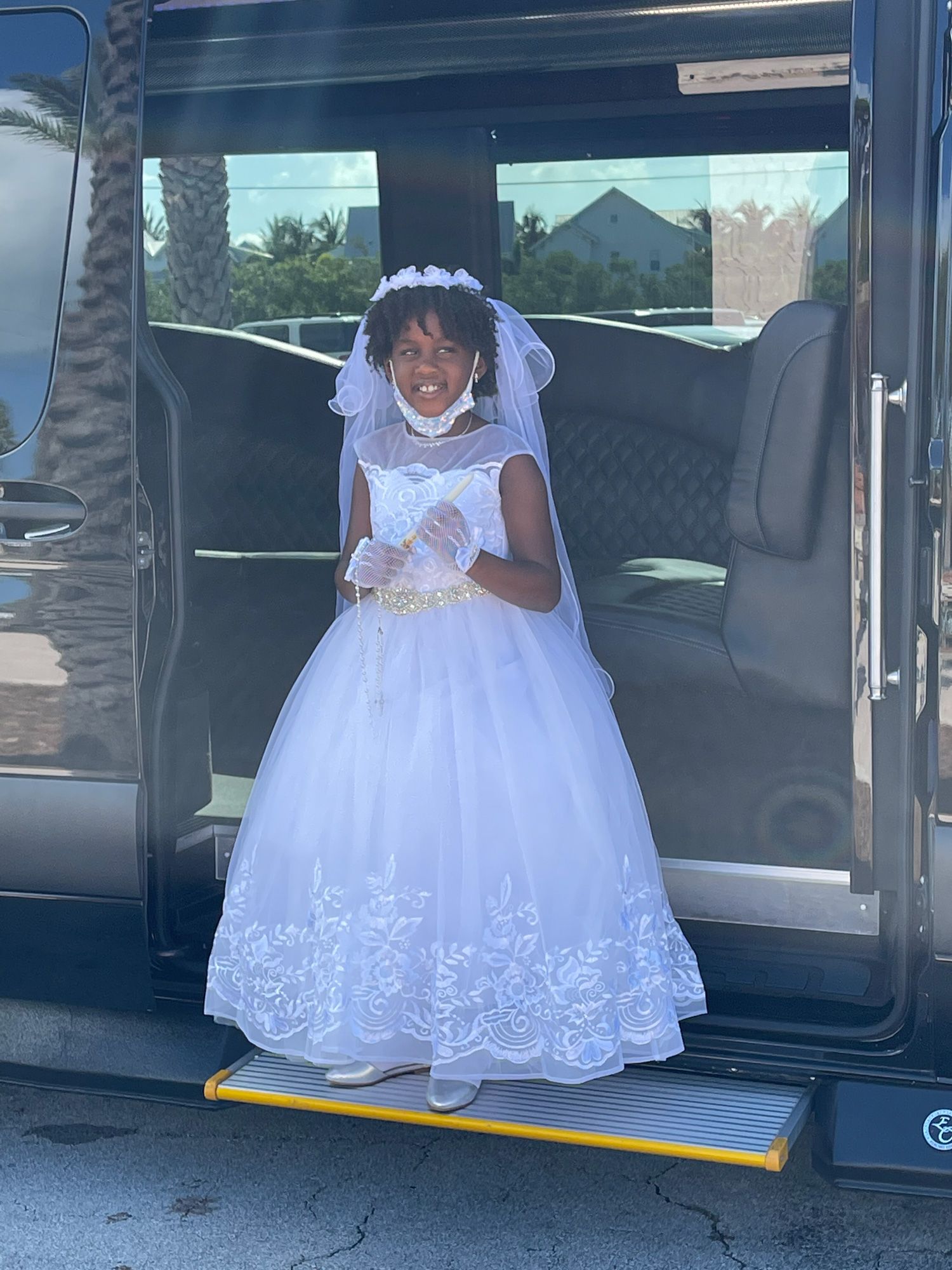 Girl in white dress and veil, smiling, standing in front of a van with an open door.