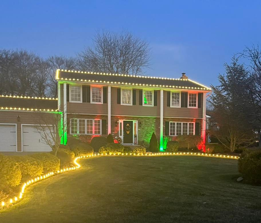 House decorated with Christmas lights: green, red, and yellow illuminate the facade and lawn at dusk.