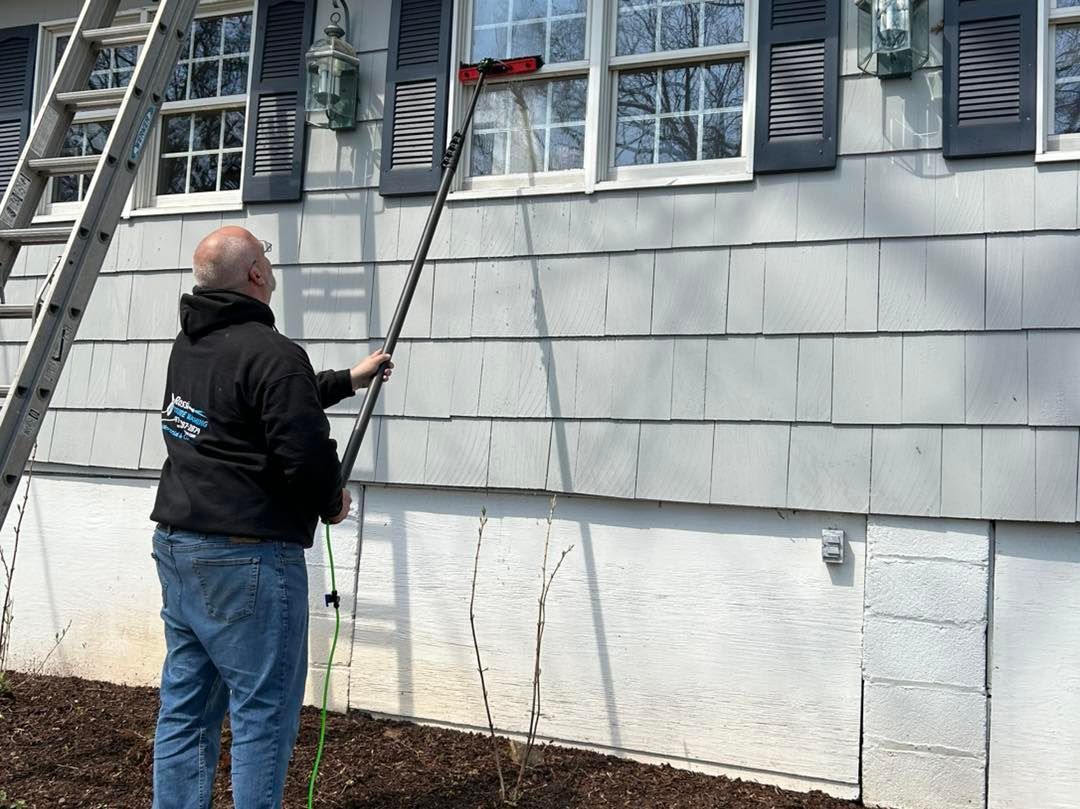 Man cleaning a window with a long brush, standing on the ground, beside a ladder.