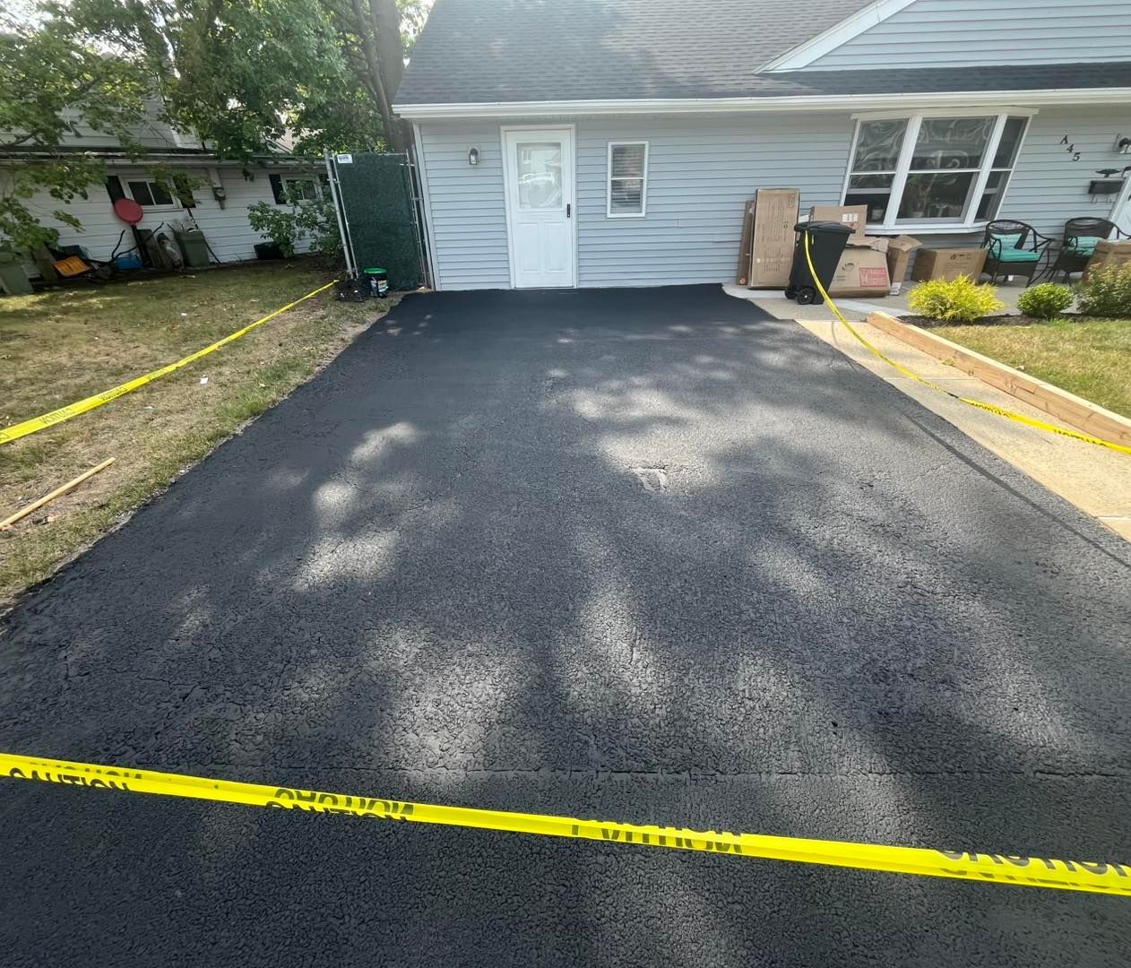 Newly paved black asphalt driveway in front of a light blue house, with yellow caution tape around the edges.